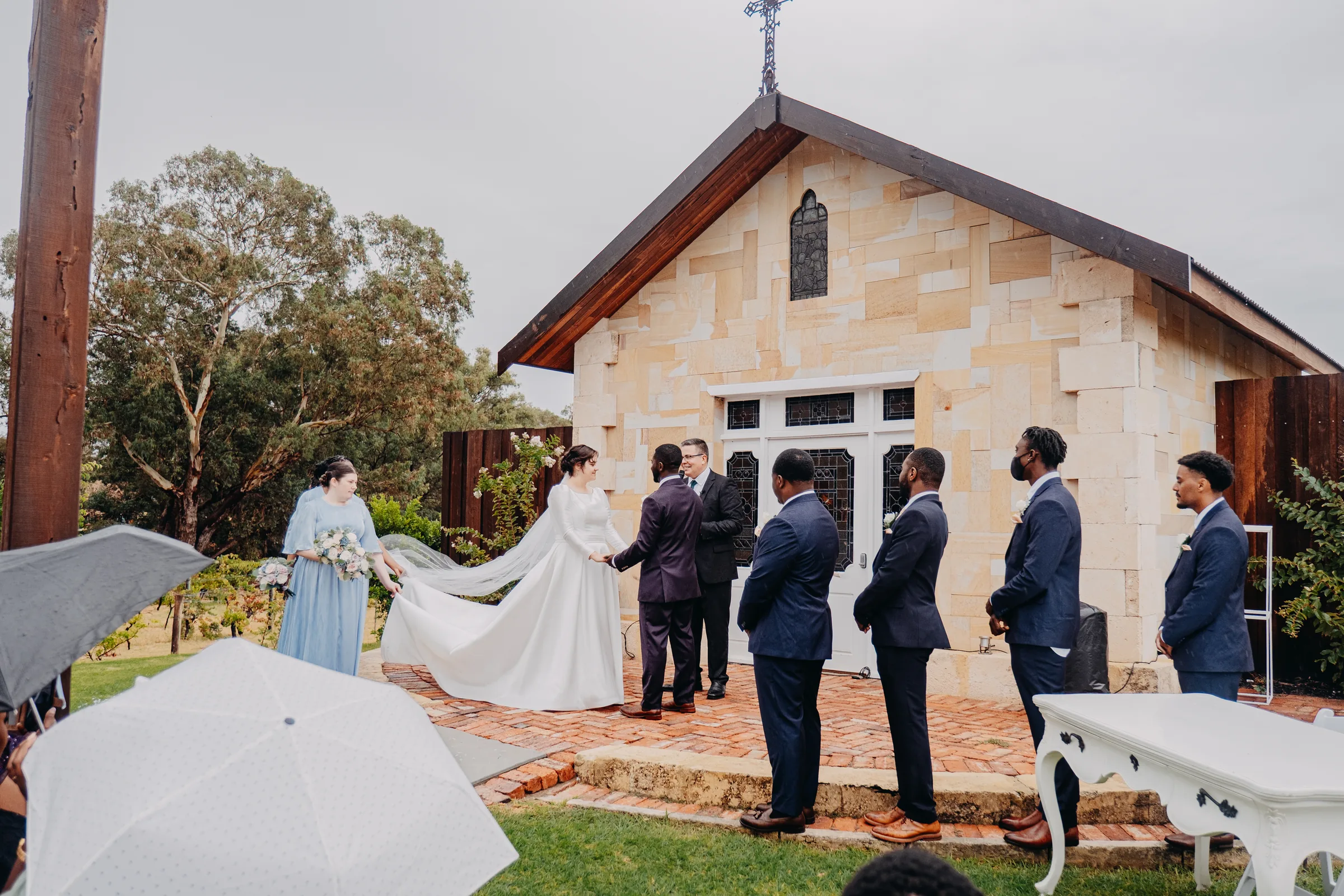 Cassandra walking down the aisle in the rain, her veil held up by a bridesmaid, toward the sandstone chapel at Chapel Farm Estate
