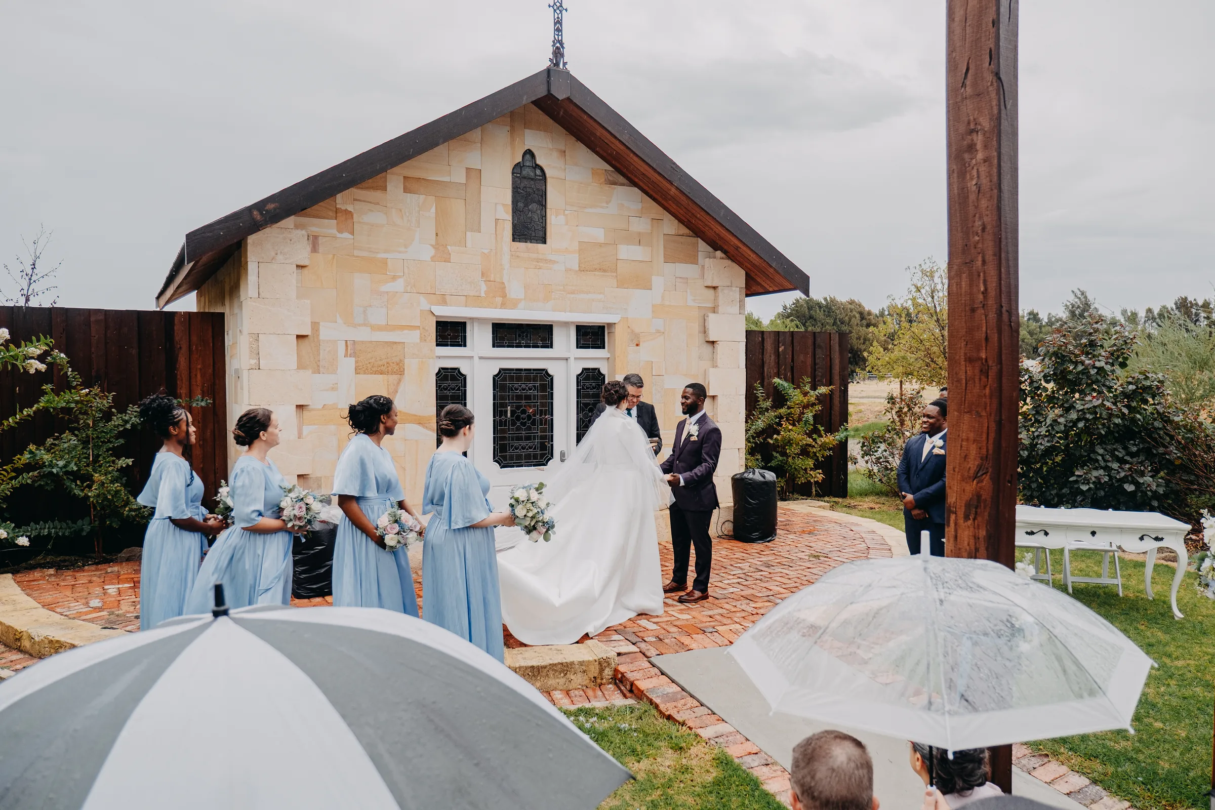 Chapel Farm Estate's sandstone chapel under grey skies with wedding guests sheltering beneath umbrellas
