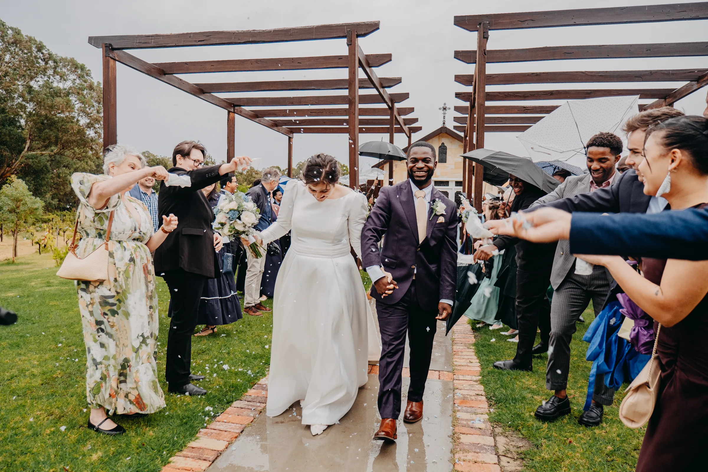Newly married Cassandra and Jonathan walking out through the rain under umbrellas as guests celebrate at Chapel Farm Estate