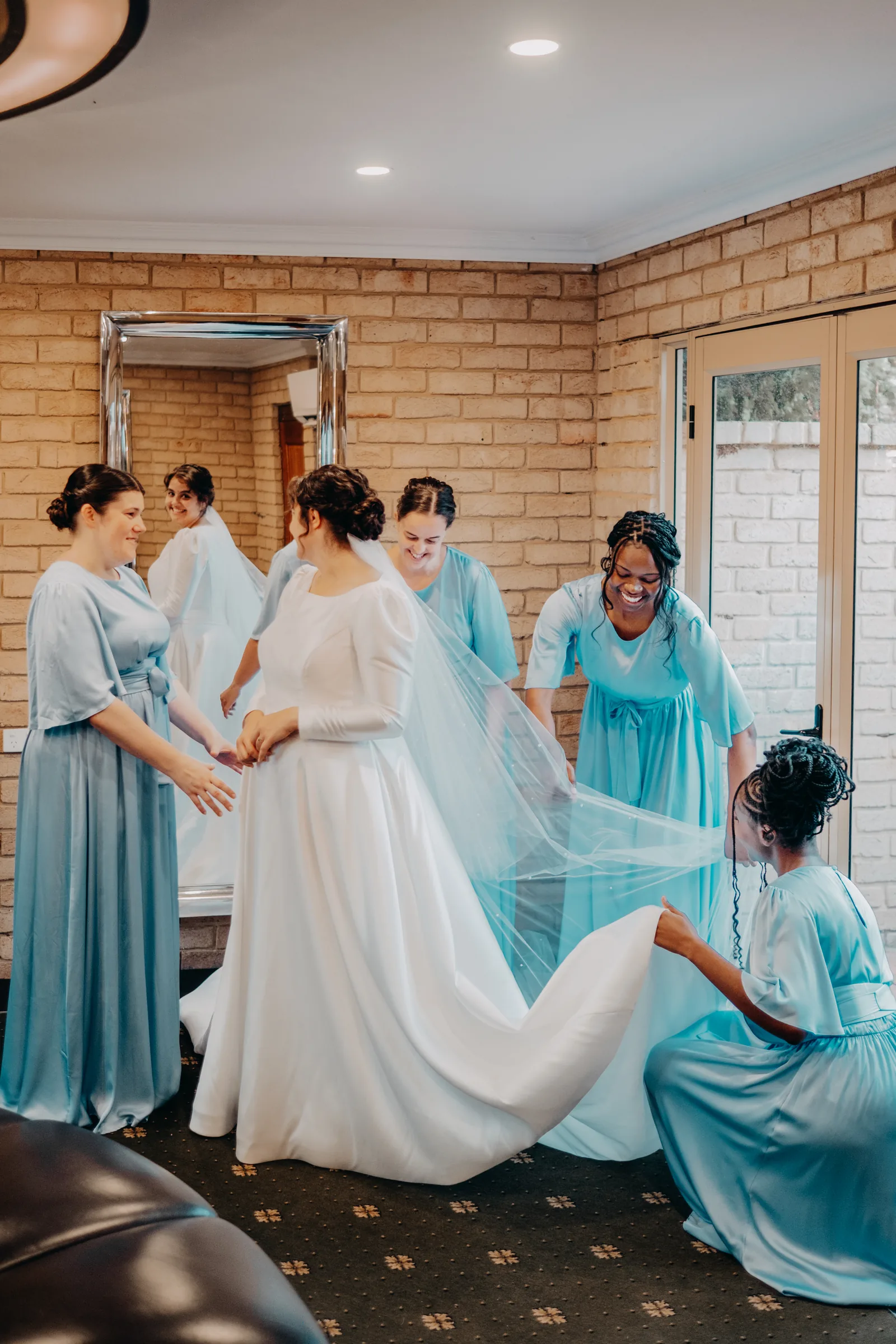 Bridesmaids in blue helping carry Cassandra's dress train through the Farm Boudoir at Chapel Farm Estate