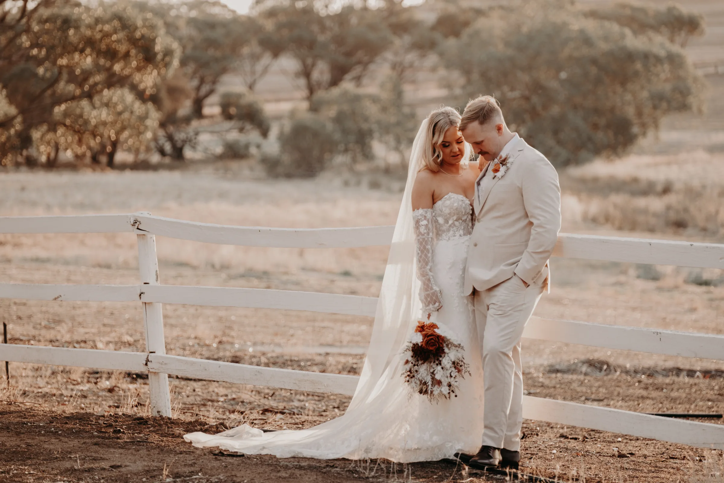Grace and Lachie standing by a timber fence with the paddocks glowing gold at sunset, Buckland Estate
