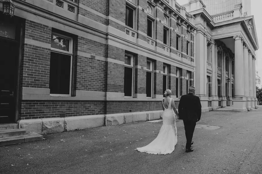Black and white photo of Chantelle and Zach walking beside the heritage Supreme Court Gardens building, Perth