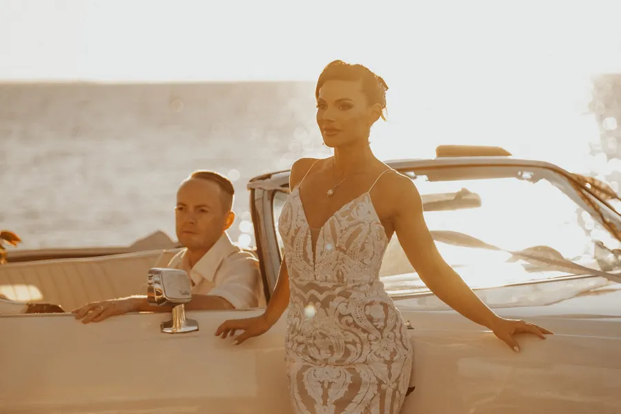 Katia and Sean leaning against their vintage Cadillac Eldorado, warm golden hour light off the water