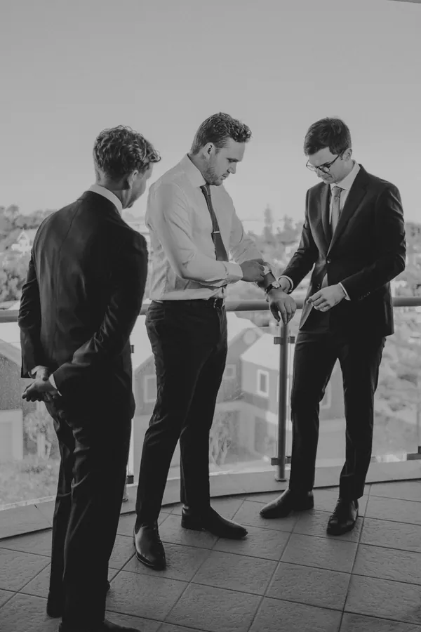 Groom and groomsmen helping each other with cufflinks and a watch while getting ready on the balcony