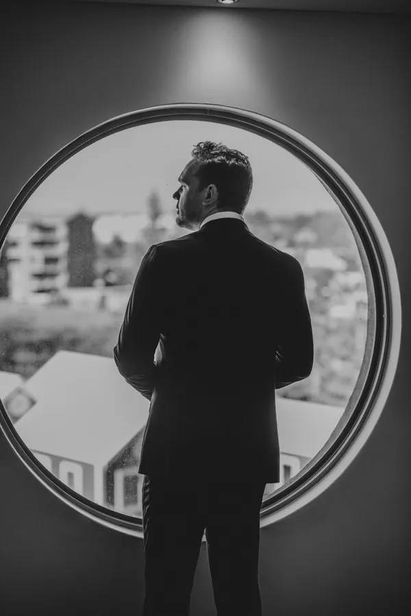 Groom looking out over Hopeland from a round window, back to camera, moments before the ceremony