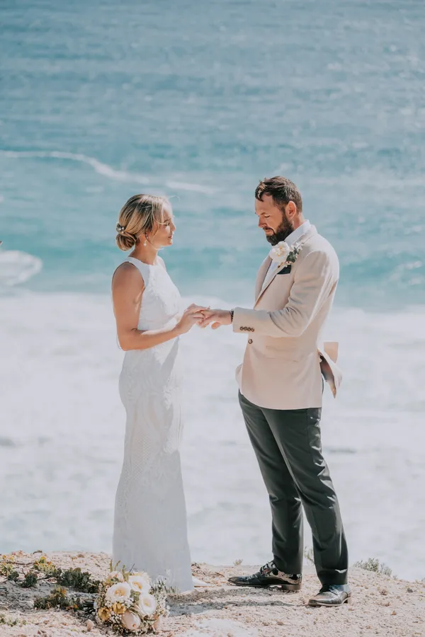 Victoria and Luck holding hands during the ring exchange, ocean blurred behind them