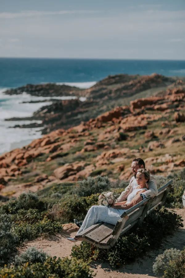 Victoria and Luck sitting close together on the bench, wrapped in each other's arms above the rocky coastline