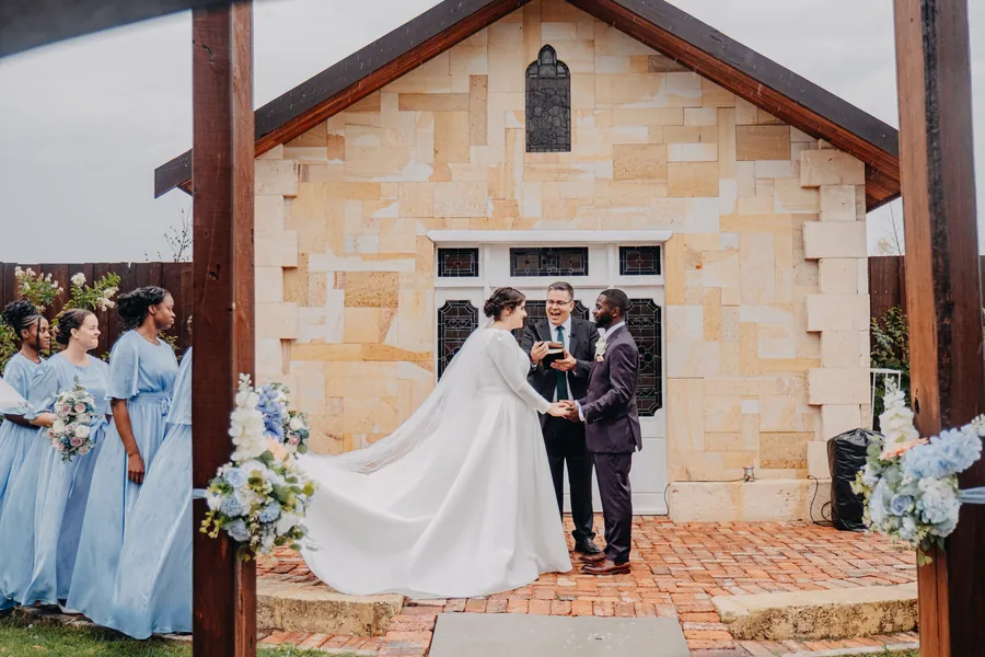 Cassandra and Jonathan at the altar in front of the sandstone chapel doors, guests holding umbrellas in the rain