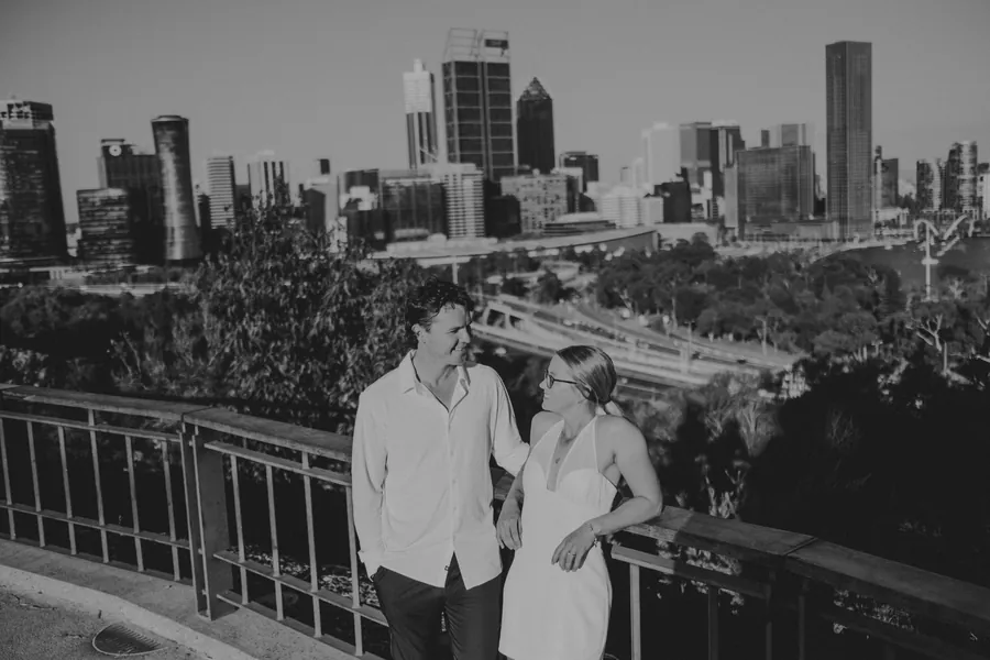 Kayla and Brett standing together at a Kings Park lookout railing with the Perth skyline behind them, black and white